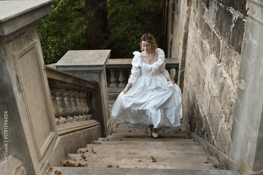 Naklejka premium full length portrait of beautiful blonde female model wearing historical white gothic wedding gown, walking down staircase of romantic fairytale castle location with stone balconies