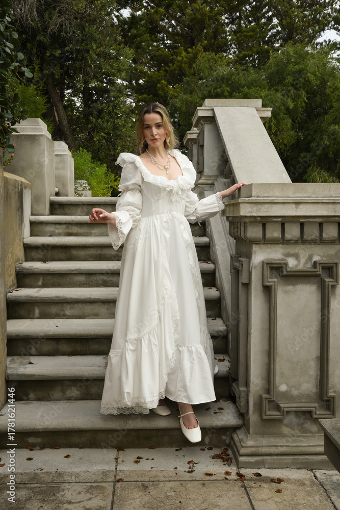 Fototapeta premium full length portrait of beautiful blonde female model wearing historical white gothic wedding gown, walking down staircase of romantic fairytale castle location with stone balconies