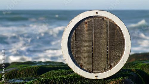 A circular wooden barrel lid, framed in white, sits on mossy rocks with the ocean as background