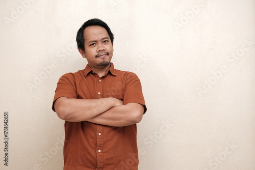 Southeast Asian man in a brown shirt standing indoors with arms crossed and a calm, confident facial expression.