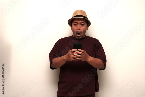 A surprised Southeast Asian man in casual clothing and hat reacting dramatically while reading something on his smartphone indoors.
