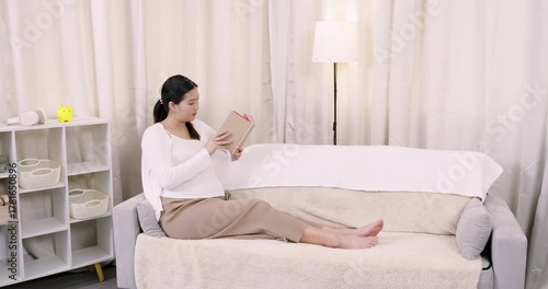 Pregnant asian woman in third trimester slowly flipping through book while relaxing on bed during peaceful afternoon, enjoying quiet selfcare moment in slowmotion to embrace maternal lifestyle