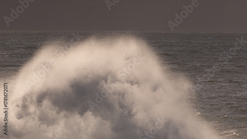 Powerful ocean wave breaking with immense white spray and foam against a dark, dramatic sea and muted horizon.