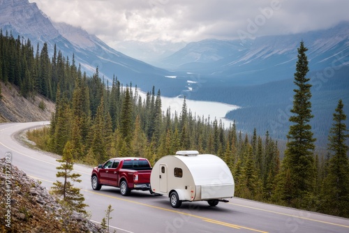 A red pickup truck tows a small white camper along a scenic mountain road beside a lake