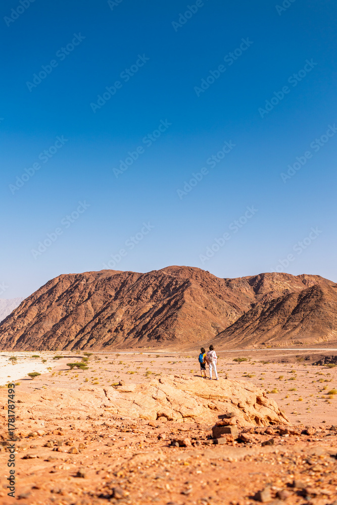 Naklejka premium A boy with a backpack and a map walks next to his mother through the desert. Family trekking. Timna National Park. The child is actively spending time.