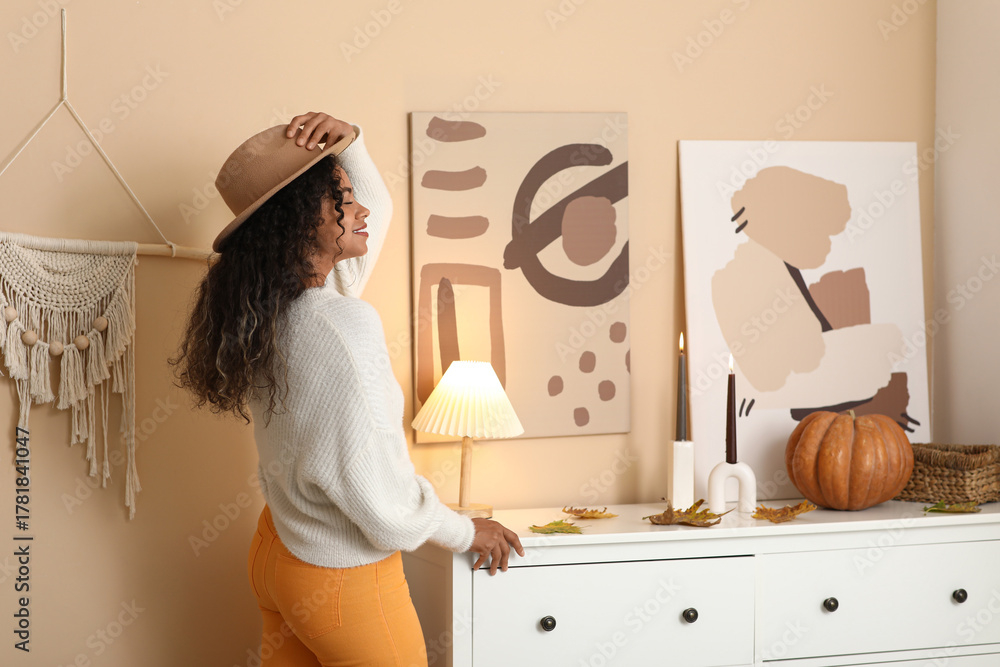 Obraz premium Beautiful young happy African-American woman in stylish outfit with autumn leaves, pumpkin and hat at home