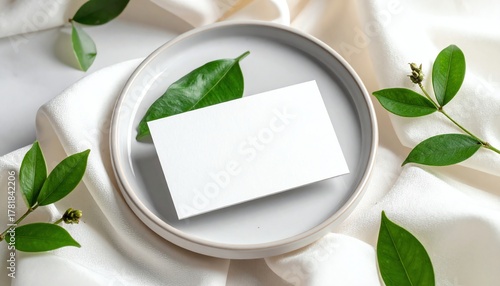 Blank card on ceramic plate surrounded by green leaves and soft white fabric, ideal for branding or elegant invitation mockup presentations.