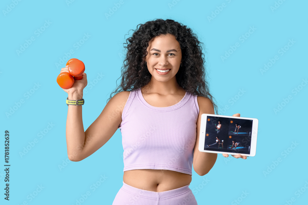 Fototapeta premium Sporty young African-American female coach with tablet computer and dumbbells on blue background. Concept of online training