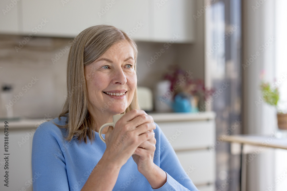Fototapeta premium Senior woman smiling warmly while holding a warm mug with both hands, enjoying a quiet moment of relaxation and contemplation in her bright home kitchen