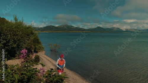 Young attractive man in shorts and jacket goes along narrow path on shore of sea bay with lush vegetation and range of mountains on horizon. Concept privacy