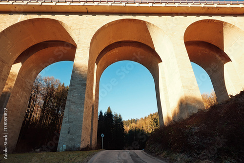 Majestic concrete bridge with large arches towering over a winding road, surrounded by lush greenery and clear blue sky, showcasing architectural beauty