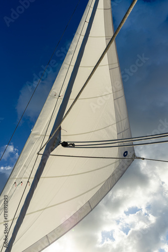 A single yacht sail catches the sunlight, glowing against a vivid blue sky scattered with soft white clouds