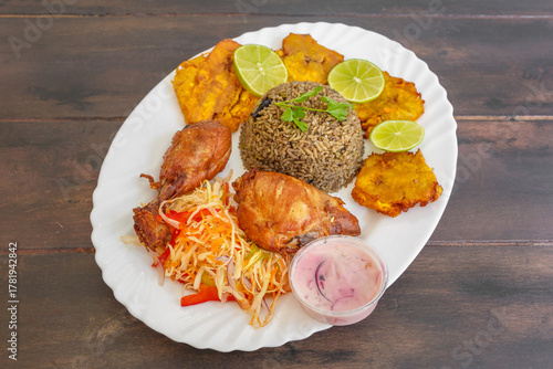 Poulet frit épicé servi avec pikliz aux poivrons, riz Djon Djon et bananes pesées frites, cuisine haïtienne traditionnelle sur une table en bois. 