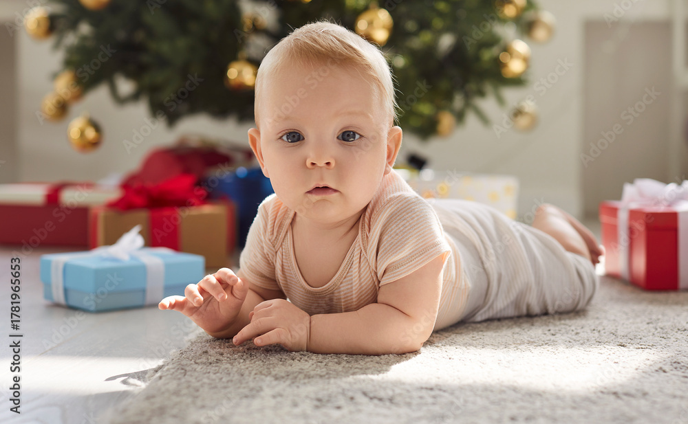 Fototapeta premium Happy small baby, first Christmas eve child on warm carpet near decorated festive tree, present boxes at home, cute little infant toddler, playing indoors upbringing, childcare and family love