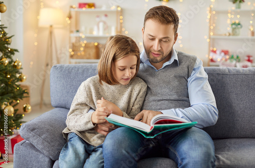 Father and son reading a book together at home during Christmas or New Year, creating a bonding experience. Shared leisure time fosters closeness, nurturing family warmth during the holiday season.