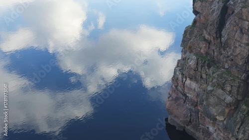 Powerful granite cliff rising over dark river water with clear reflection of the sky and white clouds.