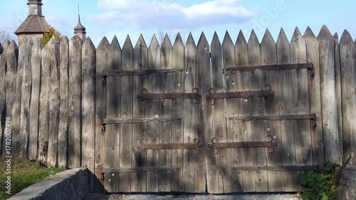 Old wooden gate or palisade with pointed stakes and heavy forged metal hinges. Historical background.