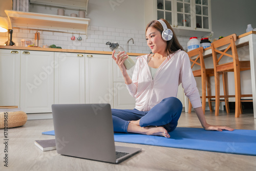 beautiful Asian woman enjoys healthy lifestyle at home wearing sporty activewear listening music through headphones drinking nutritious protein milkshake yoga relaxed atmosphere reflects modern life