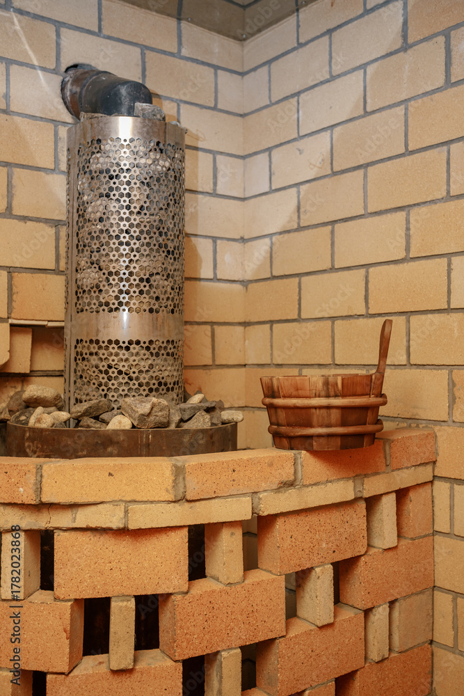 Fototapeta premium Vertical shot of a brick-covered sauna stove with hot stones, with a water ladle placed on the edge. A warm brick wall in the background creates a rustic and traditional steam room atmosphere.