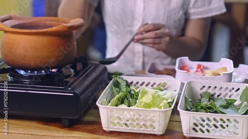Woman Cooking in Traditional Style with Clay Pot on Stove Surrounded by Fresh Vegetables and Herbs on Wooden Table in Cozy Kitchen Setting