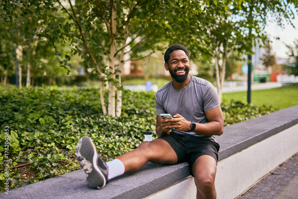 Fototapeta premium Smiling Man in Workout Gear Using Phone Outdoors While Relaxing on Park Ledge
