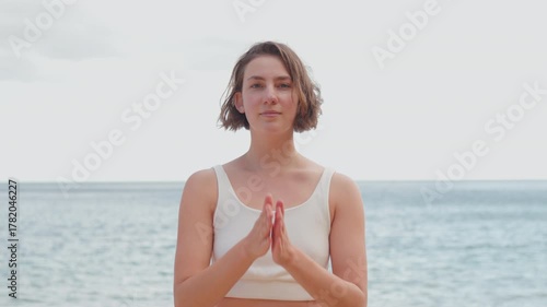 Young woman with short hair in prayer pose with hands together smiling and meditating by sea representing inner calm and positive energy during daytime