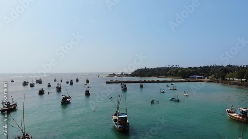 Stunning aerial perspective of traditional fishing boats floating on the calm turquoise water of a tranquil harbor, with a scenic coastline featuring a jetty and lush green palm trees