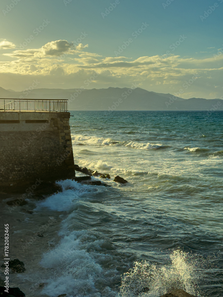 Fototapeta premium Waves crashing against a stone pier at sunset near a coastal town with mountains in the backdrop