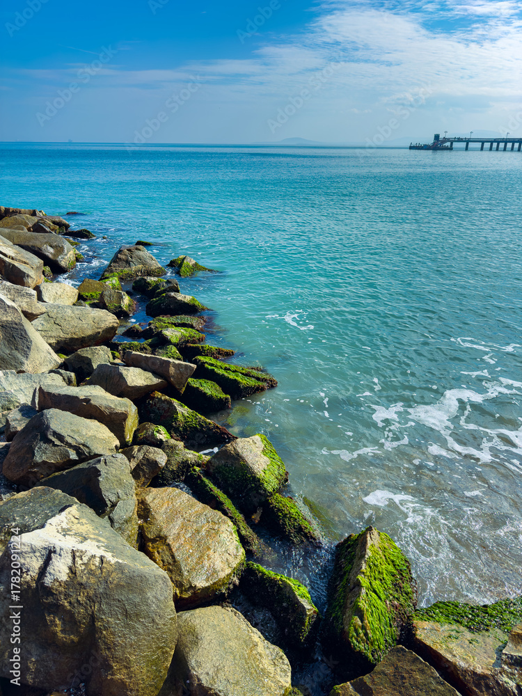 Fototapeta premium Vibrant coastal scene showcasing rocky shoreline and tranquil waters of the seaside at midday with a distant pier under a bright blue sky
