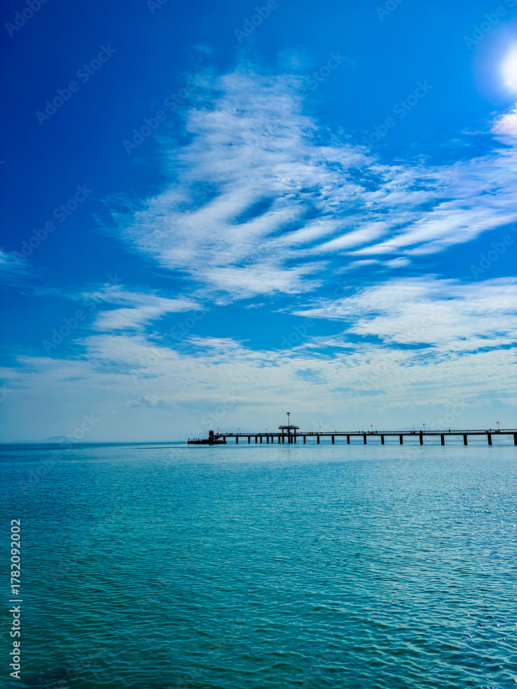 Fototapeta premium Sunlight dances on tranquil waters at a serene pier surrounded by a bright blue sky with soft clouds as a boat glides by