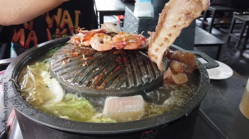 Close-up of Thai-style barbecue grill pan, known as Moo Kratha, with pork belly, shrimp, and vegetables cooking on a hot grill and soup. Popular Asian dining experience in Thailand.