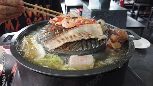 Close-up of Thai-style barbecue grill pan, known as Moo Kratha, with pork belly, shrimp, and vegetables cooking on a hot grill and soup. Popular Asian dining experience in Thailand.