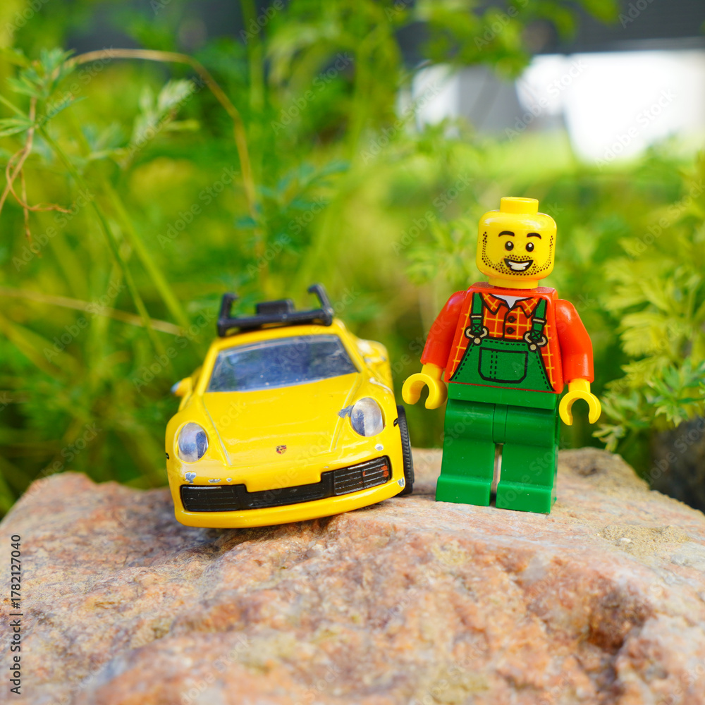 Obraz premium Lego figure with a yellow toy car on a rock in front of green foliage