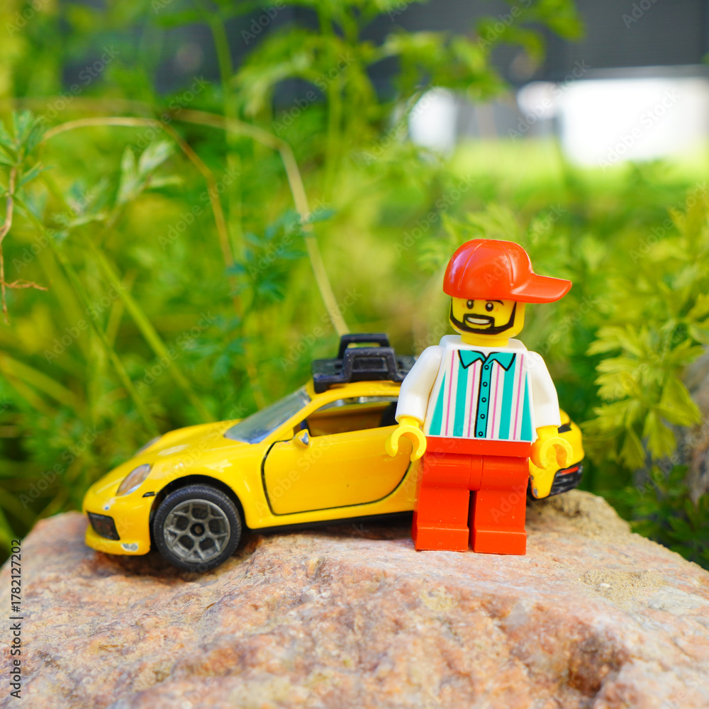 Obraz premium Lego figure with yellow toy car on a rock in front of green foliage