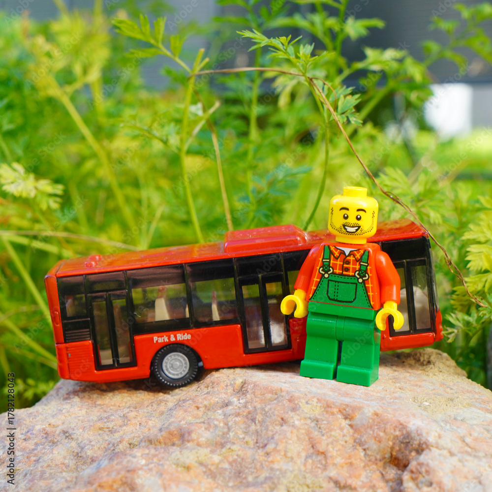 Naklejka premium Lego minifigure with a red bus on a rock in front of green foliage