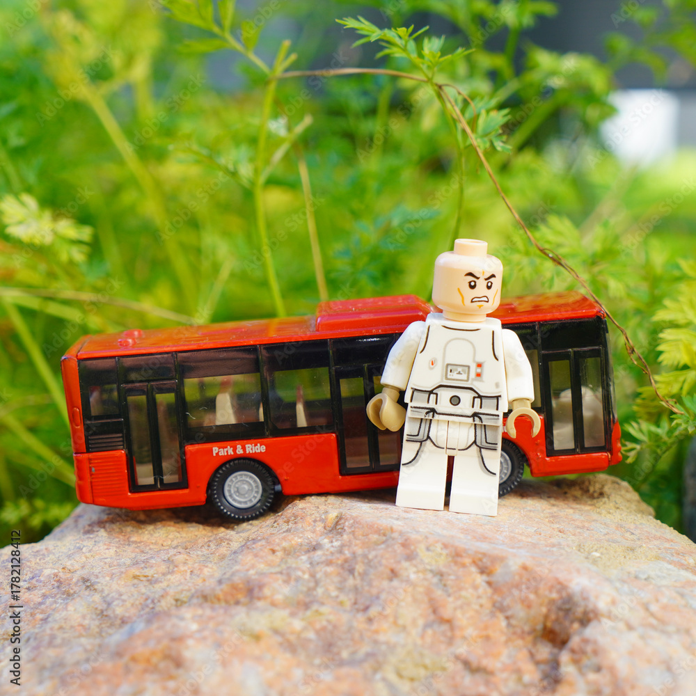 Naklejka premium Lego figure stands near a red toy bus on a rock with green foliage background