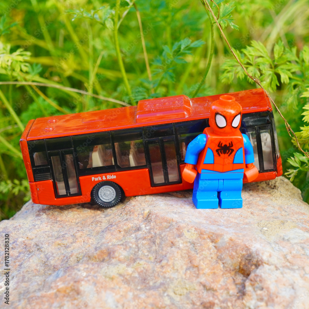 Naklejka premium Spider-Man toy figure stands next to a red toy bus on a rock outdoors