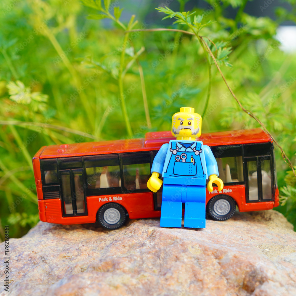 Naklejka premium Lego bus driver figure stands next to a red toy bus on a rock