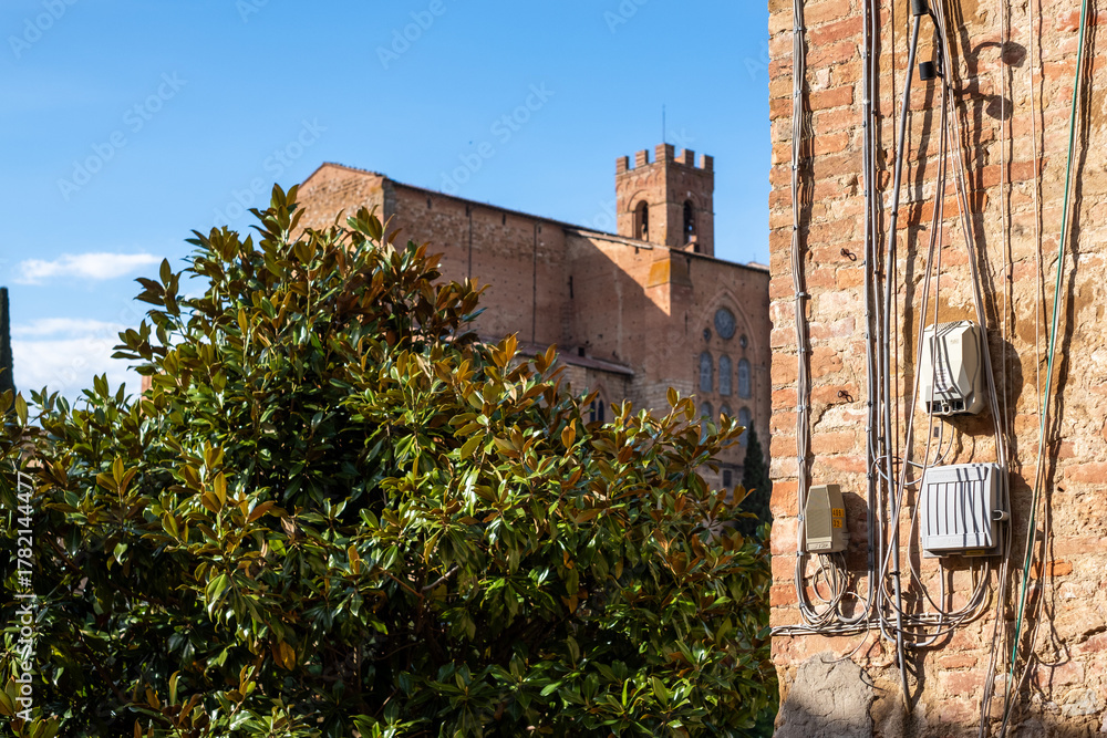 Obraz premium Electrical installation on a brick wall with visible wires and safety features, surrounded by greenery and historical architecture, showcasing urban infrastructure and design concepts