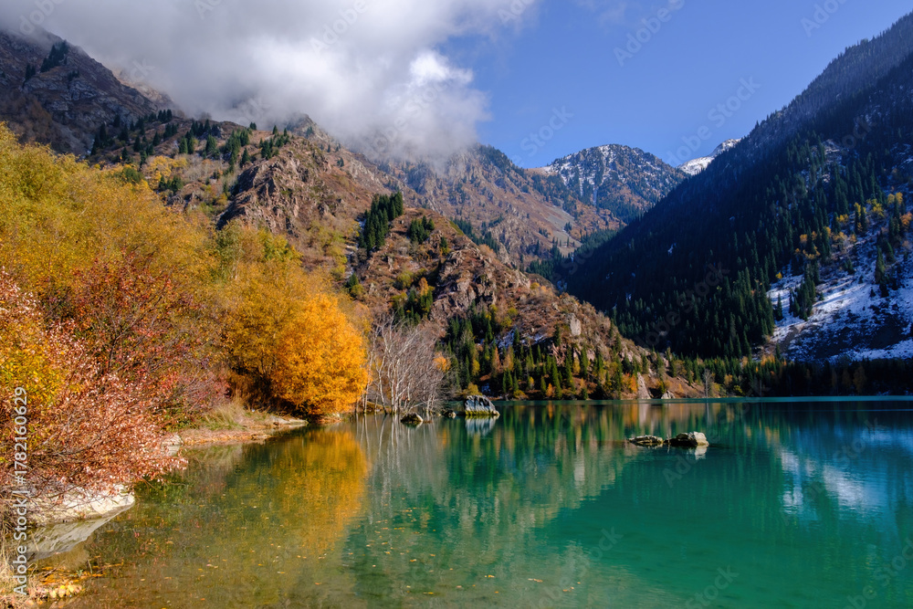 Naklejka premium Lake Issyk in autumn colors. The turquoise surface of the water reflects the snowy peaks and golden forests, creating a mesmerizing landscape.