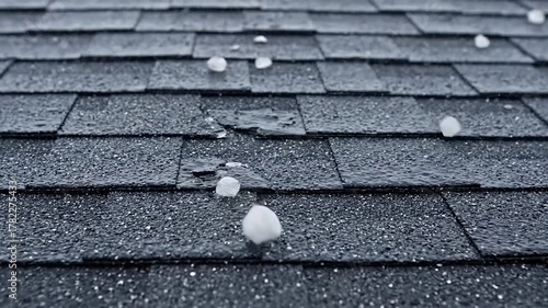 Close up view of large hailstones falling and bouncing on asphalt roof shingles during a severe weather storm, causing damage