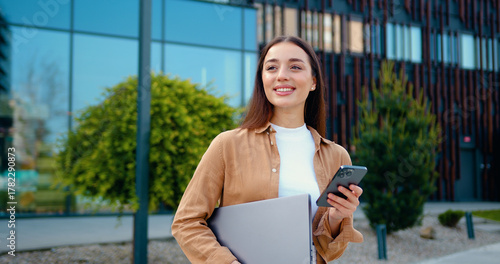 Portrait of Young Caucasian business woman in casual outfit use smartphone and holding laptop in hand while walking at front of modern Office Building. Business and people concepts