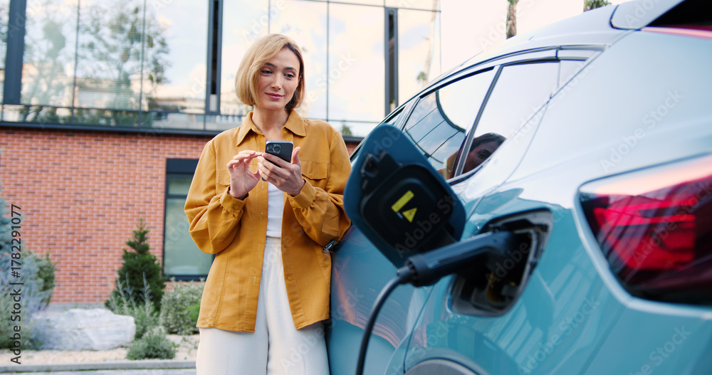 Obraz premium Woman using smartphone app while standing next to her electric car charging at a portable charging station on a city street.