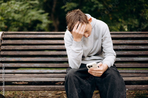 Teen boy sitting on park bench shocked looking at smartphone screen.