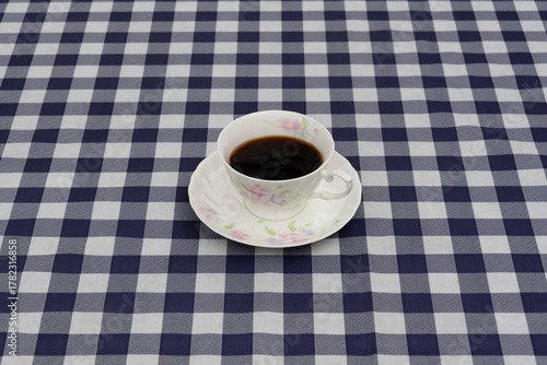 Top view of a porcelain coffee cup with floral pattern and espresso, on a matching saucer. Set on a large, navy blue and white plaid tablecloth. Symbolizes breakfast, break, and home.