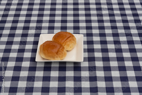 Two sweet rolls on a white square plate, centered on a navy blue and white plaid tablecloth. Top view. Symbolizes breakfast, bakery, snack, and simple comfort food.