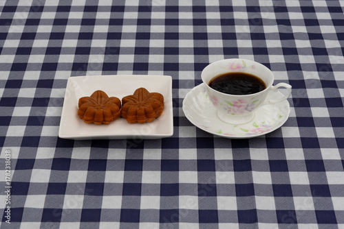 Top view of a coffee cup and a plate with two maple leaf-shaped pastries on a navy blue and white plaid tablecloth. Ideal for breakfast or autumn snack concept.