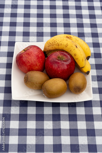 Close-up of a white square plate with fresh fruit (apples, kiwis, bananas) on a navy blue and white plaid tablecloth. Vertical orientation. Ideal for healthy snack and diet concepts.