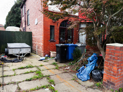 Cluttered alley beside a red brick house in Manchester showing garbage, bins, and overgrowth