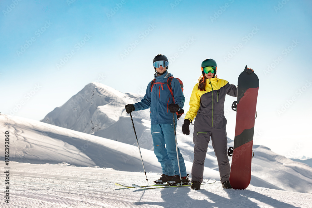 Obraz premium Couple skier and snowboarder stand at the top of a ski resort slope on sunny day with snow-capped peaks in the background.